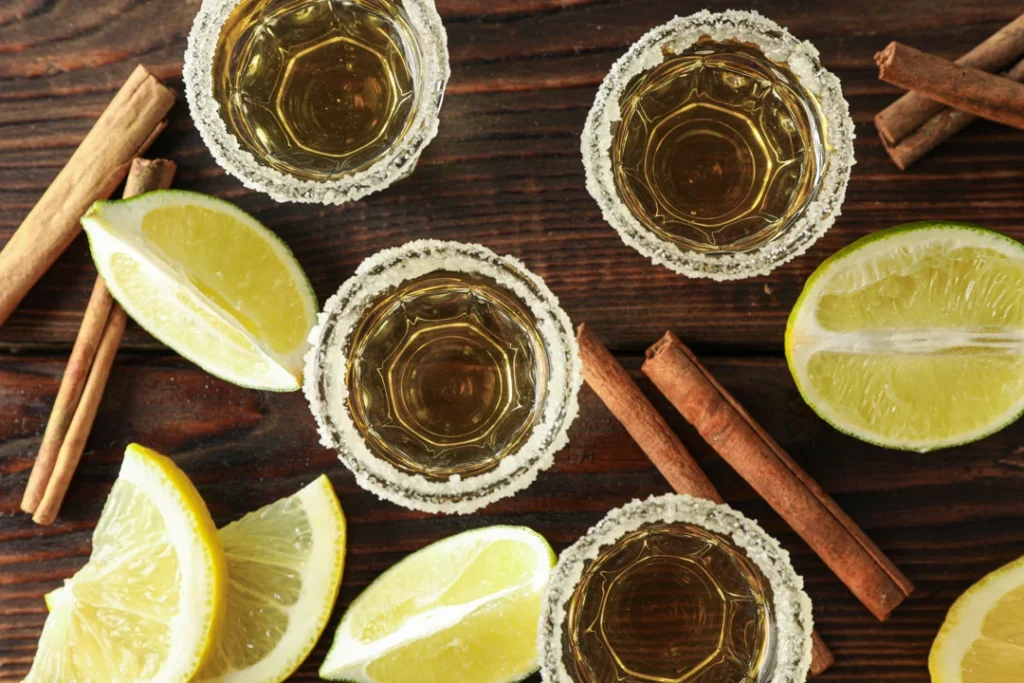 Tequila shots with salt, lime slices and cinnamon on wood table, top view